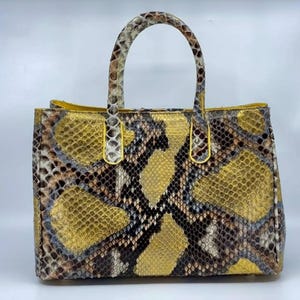 May include: A rectangular handbag with a top handle, crafted from a snakeskin pattern. The bag features a mix of brown, black, and gold tones, with a yellow interior. The handle and trim are a lighter shade, creating a striking contrast.