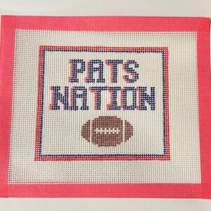 May include: A square needlepoint canvas featuring a white mesh background with a bright pink fabric border. The design includes the text PATS NATION in dark blue with red shadowing, positioned above a brown football graphic with white stitching.
