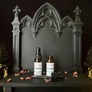 May include: Two small brown glass bottles with black droppers are displayed on a black shelf. The bottles have white labels with red text. The shelf is part of a gothic-style structure with a fleur-de-lis design. Dried rose petals are scattered around the bottles.
