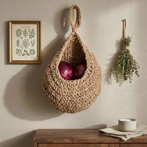 May include: A woven jute hanging basket with two red onions inside. A framed botanical print and a bundle of herbs hang on the wall. A small bowl of salt and a folded linen napkin sit on a wooden surface.