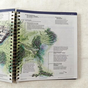 May include: Open spiral-bound notebook with detailed map illustrations. The pages display a green and tan topographical map with roads, buildings, and text. The notebook has a dark blue cover and gold spiral binding.