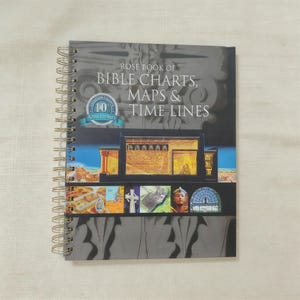 Könnte beinhalten: Ein spiralgebundenes Buch mit dem Titel "Rose Book of Bible Charts, Maps & Time Lines" mit einem dunkelgrauen Einband. Der Einband zeigt architektonische Illustrationen und ein 10-jähriges Jubiläumssiegel. Die Spiralbindung des Buches ist goldfarben.