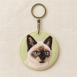 Può includere: Portachiavi ricamato con un motivo di gatto siamese. Il gatto ha occhi blu, orecchie e segni sul viso marrone scuro e un corpo color crema. Il portachiavi ha uno sfondo rotondo verde chiaro con un anello e una chiusura in metallo color oro.