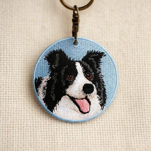 May include: Embroidered keychain featuring a Border Collie dog. The dog's face is detailed with black and white fur, brown eyes, and a pink tongue, set against a light blue circular background. A metal keyring is attached at the top.