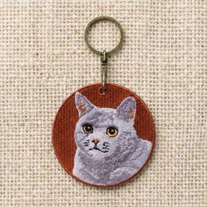 Può includere: Un portachiavi rotondo ricamato con un gatto grigio con occhi gialli su sfondo color ruggine. Il ritratto del gatto è dettagliato con pelo e baffi cuciti. Il portachiavi ha un anello e una chiusura color bronzo.