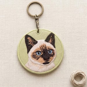 Può includere: Portachiavi ricamato raffigurante un gatto siamese con occhi blu e segni marroni. Il portachiavi rotondo ha un bordo verde chiaro e un anello e una chiusura in metallo color bronzo.