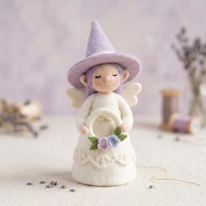 May include: A handcrafted felt angel doll wearing a lavender witch hat and holding a floral wreath. The doll has white wings, a white dress, and closed eyes. The background is a soft purple, with lavender sprigs and a spool of thread.