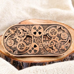 Puede incluir: Hebilla de cinturón de estilo steampunk en color cobre con un intrincado diseño de engranajes, ruedas dentadas y los números "2018" y "12". La hebilla ovalada descansa sobre una superficie de madera.