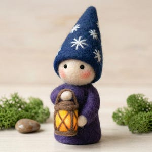 May include: A handmade felt gnome doll wearing a dark blue hat with white snowflake designs and a purple tunic. The gnome holds a small, lit lantern with an orange glow. The doll has rosy cheeks and a neutral expression.