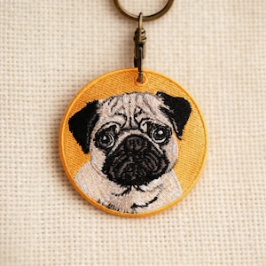 May include: Embroidered round keychain featuring a pug's face. The pug has a tan and black coat, with large, dark eyes. The keychain's background is a bright yellow, and it has a metal clasp and ring.