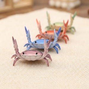 Könnte beinhalten: Eine Sammlung von Miniatur-Krabbenfiguren in verschiedenen Farben, darunter Rosa, Blau, Orange und Grün. Jede Krabbe hat detaillierte Scheren und einen Panzer, angeordnet auf einer strukturierten Oberfläche. Diese dekorativen Figuren eignen sich für Küsten-Dekorationen.