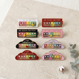May include: A collection of enamel pins with the phrases "MY SOCIAL BATTERY" and "MOODTRACKER". The pins feature a battery or cloud design with a mood indicator. Colors include white, red, black, pink, and gold.