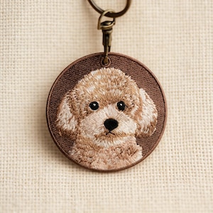 Peut inclure: Porte-clés brodé représentant un chien caniche de couleur marron clair. Le visage du chien est au centre d'un cadre circulaire marron foncé. Un fermoir de couleur bronze est attaché. La fourrure du chien est détaillée avec du fil beige et marron clair.
