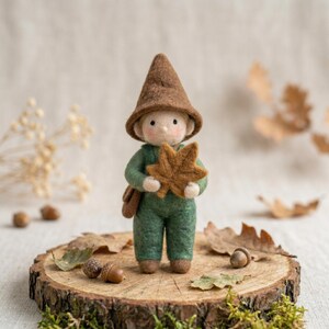 May include: A handmade felt gnome doll in a green jumpsuit and a brown pointed hat. The gnome holds a brown felt leaf and has a small brown satchel. The doll is standing on a wooden slice with acorns and leaves.