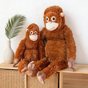 May include: Two plush orangutan stuffed animals in varying sizes. The larger one sits beside the smaller one. Both have brown fur, white faces, and large, expressive eyes. They are positioned on a wooden surface.