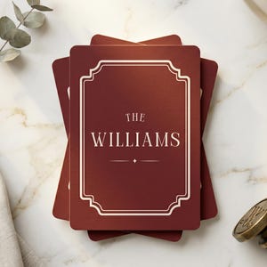 Op de afbeelding: Een stapel rechthoekige donkerrode onderzetters met een crèmekleurige decoratieve rand en de tekst "THE WILLIAMS" in het midden gedrukt. Het ontwerp bevat een klein vierpuntig stersymbool onder de familienaam.