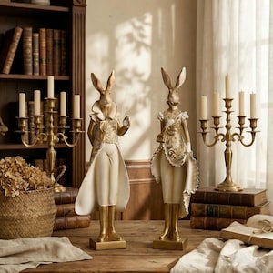 Op de afbeelding: Twee decoratieve konijnenbeeldjes, een met een zakhorloge, de ander met een kleine tas. Beide dragen crème- en gouden outfits met ingewikkelde details. Ze staan op gouden sokkels, met een vintage kandelaar en boeken op de achtergrond.