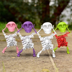 May include: Four colorful skeleton figurines. Each skeleton has a different colored skull: pink, purple, white, and green. The skeletons are white with matching colored limbs. They are standing on a gray surface.