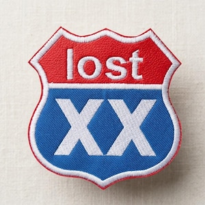 Op de afbeelding: Een schildvormige patch met een rood bovenstuk en een blauw onderste gedeelte. Het woord "lost" is in het wit geborduurd op het rode gedeelte en de letters "XX" zijn in het wit geborduurd op het blauwe gedeelte. De patch heeft een witte rand.