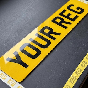 Puede incluir: Una placa de matrícula rectangular amarilla con superficie reflectante y letras negras que dicen YOUR REG. La placa presenta un patrón repetitivo del logotipo AP a lo largo de los bordes superior e inferior, con un diseño claro y contrastado.