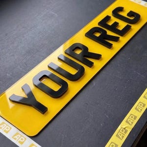 May include: A rectangular vehicle license plate featuring a bright yellow reflective background and raised, three-dimensional black lettering that spells out the text YOUR REG. The plate has a smooth, glossy finish with clean, sharp edges.