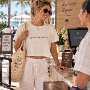 May include: A cream-colored cropped t-shirt with the black text 'LATE CHECK OUT PLEASE' and a palm tree icon, paired with white drawstring pants and a beige woven tote bag featuring the black text 'Same Shift Different View' with a heart.