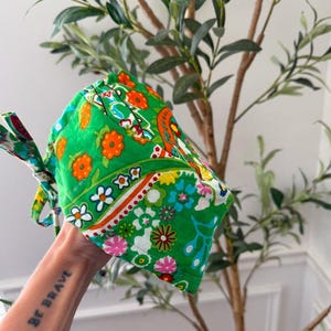 May include: A green patterned scrub cap with orange, white, and blue floral designs. The cap has ties for securing and is made of a soft fabric. The cap is designed for medical professionals.