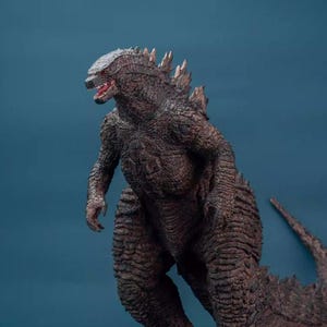 May include: A detailed brown Godzilla figure with textured skin and dorsal fins. The figure has an open mouth, revealing red details, and is posed on a blue background. The tail extends to the right.