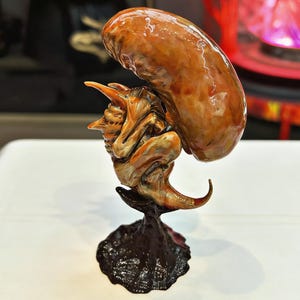 May include: A detailed sculpture of a fantastical creature with a large, brown, oblong form balanced on its back. The creature is primarily orange and brown, with a textured appearance and a dark, textured base.