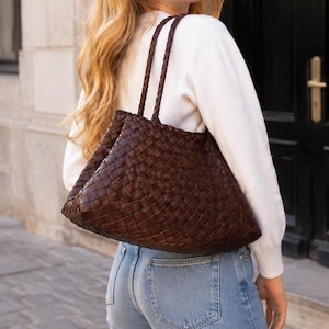 May include: A dark brown woven leather tote bag featuring a large, intricate basket-weave pattern throughout the body. The bag includes two thin, braided leather shoulder straps attached to the top edge, providing a structured yet flexible design.