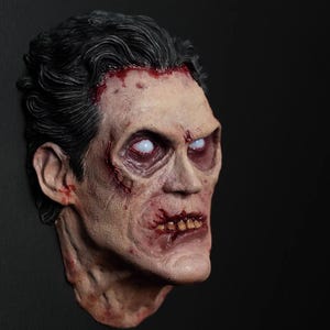 May include: A detailed zombie head sculpture with pale skin, dark hair, and blood-red accents. The eyes are white, and the mouth is open, revealing teeth. The sculpture is set against a black background.