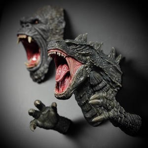 May include: Two detailed wall sculptures of iconic movie monsters. One depicts a roaring King Kong head, the other a Godzilla head with open mouth and sharp teeth. The sculptures are dark grey with red and pink interior details.