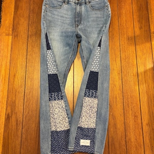 May include: Light blue denim jeans with patchwork panels down the sides. The patchwork features various blue and white floral and geometric patterns. The jeans are displayed on a wooden surface.