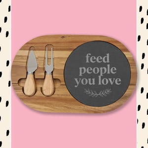 May include: A wooden cheese board with a slate insert. The board includes a cheese knife and fork with wooden handles. The slate insert has the text "feed people you love" in white lettering with a small leaf design.