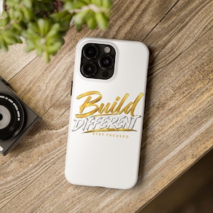 Peut inclure: Une coque de smartphone blanche avec le texte doré et noir Build Different et le texte doré plus petit Stay Focused. La coque présente une découpe pour un système de caméra à trois objectifs sur le côté supérieur gauche, au design épuré.