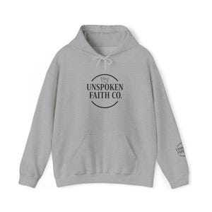 Puede incluir: Una sudadera con capucha gris claro con capucha con cordón y bolsillo delantero. La parte delantera de la sudadera con capucha presenta un logotipo circular con el texto "UNSPOKEN FAITH CO." y un pequeño emblema. La manga tiene el texto "UNSPOKEN FAITH".