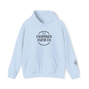 Könnte beinhalten: Ein hellblauer Kapuzenpullover mit einer Kängurutasche auf der Vorderseite und langen Ärmeln. Auf der Brust befindet sich ein schwarzes kreisförmiges Logo mit einer Taube und dem Text "UNSPOKEN FAITH CO.". Ein kleineres Logo ist am linken Ärmel.