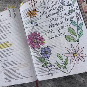 Puede incluir: Una Biblia abierta con caligraf&iacute;a manuscrita que dice The Lord gives His people strength. The Lord blesses them with peace. PSALM 29:11. La p&aacute;gina presenta flores dibujadas a mano y coloreadas en rosa, morado y naranja con hojas verdes.