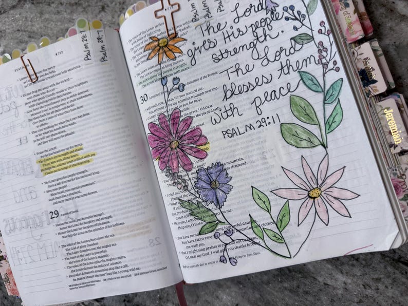Puede incluir: Una Biblia abierta con caligraf&iacute;a manuscrita que dice The Lord gives His people strength. The Lord blesses them with peace. PSALM 29:11. La p&aacute;gina presenta flores dibujadas a mano y coloreadas en rosa, morado y naranja con hojas verdes.