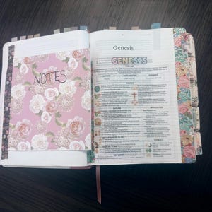 Puede incluir: Un libro abierto con una inserci&oacute;n de notas con estampado floral en la p&aacute;gina izquierda que presenta la palabra NOTES. La p&aacute;gina derecha muestra el libro del G&eacute;nesis con texto impreso, encabezados coloridos y pesta&ntilde;as florales en el borde.