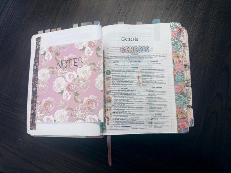 Puede incluir: Un libro abierto con una inserci&oacute;n de notas con estampado floral en la p&aacute;gina izquierda que presenta la palabra NOTES. La p&aacute;gina derecha muestra el libro del G&eacute;nesis con texto impreso, encabezados coloridos y pesta&ntilde;as florales en el borde.
