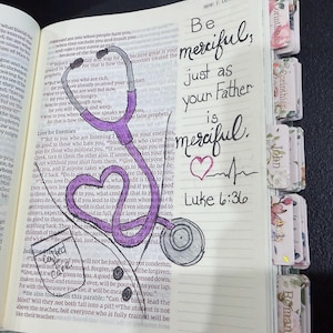 Puede incluir: Un libro abierto con texto impreso que presenta un estetoscopio morado dibujado a mano con forma de coraz&oacute;n. La p&aacute;gina incluye el texto escrito a mano: Be merciful, just as your Father is merciful. Luke 6:36, un peque&ntilde;o coraz&oacute;n y una l&iacute;nea de pulso. Tiene pesta&ntilde;as florales.