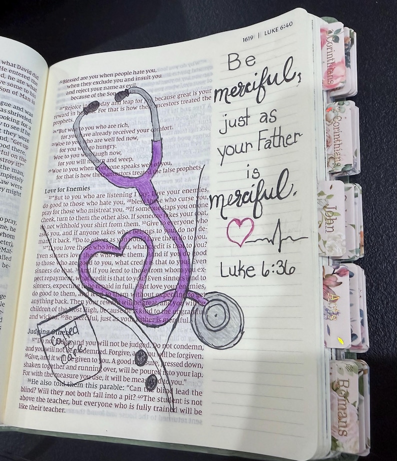 Puede incluir: Un libro abierto con texto impreso que presenta un estetoscopio morado dibujado a mano con forma de coraz&oacute;n. La p&aacute;gina incluye el texto escrito a mano: Be merciful, just as your Father is merciful. Luke 6:36, un peque&ntilde;o coraz&oacute;n y una l&iacute;nea de pulso. Tiene pesta&ntilde;as florales.