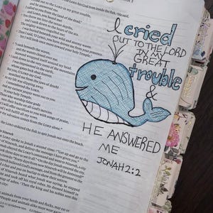 Puede incluir: Una p&aacute;gina de Biblia abierta con una ilustraci&oacute;n de ballena azul dibujada a mano y letras en tinta negra que dicen I cried out to the Lord in my great trouble and he answered me Jonah 2:2. Un clip dorado en forma de coraz&oacute;n est&aacute; sujeto arriba.