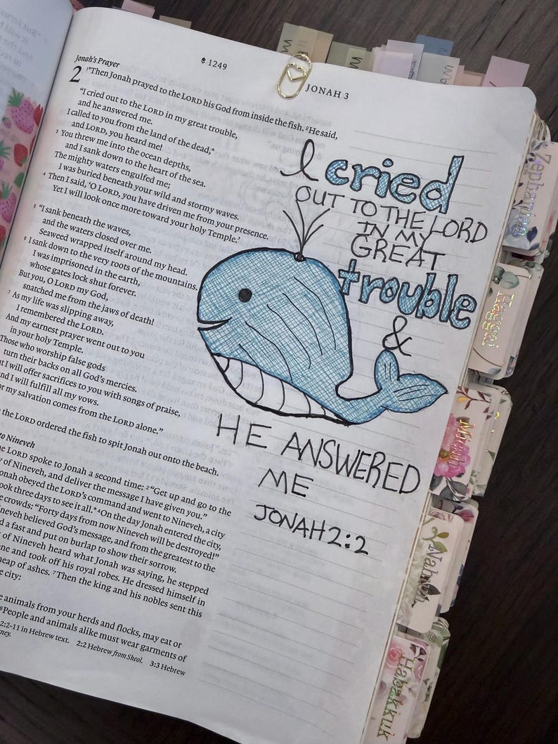 Puede incluir: Una p&aacute;gina de Biblia abierta con una ilustraci&oacute;n de ballena azul dibujada a mano y letras en tinta negra que dicen I cried out to the Lord in my great trouble and he answered me Jonah 2:2. Un clip dorado en forma de coraz&oacute;n est&aacute; sujeto arriba.