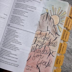 Puede incluir: Una p&aacute;gina de Biblia abierta con el Salmo 63 y una ilustraci&oacute;n dibujada a mano de una monta&ntilde;a y un sol. El texto O God, my soul thirsts for you! est&aacute; escrito en tinta negra sobre una base de acuarela. Incluye pesta&ntilde;as amarillas y un clip de coraz&oacute;n.
