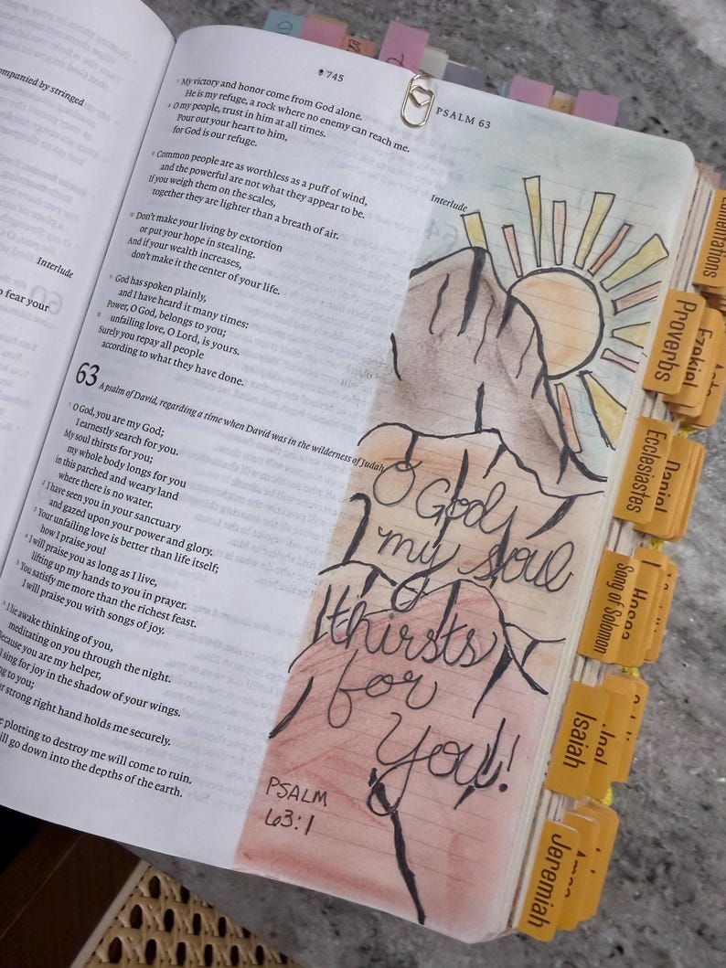 Puede incluir: Una p&aacute;gina de Biblia abierta con el Salmo 63 y una ilustraci&oacute;n dibujada a mano de una monta&ntilde;a y un sol. El texto O God, my soul thirsts for you! est&aacute; escrito en tinta negra sobre una base de acuarela. Incluye pesta&ntilde;as amarillas y un clip de coraz&oacute;n.