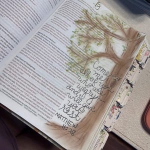 Puede incluir: Una Biblia abierta con texto impreso y una ilustraci&oacute;n en acuarela de un &aacute;rbol. La caligraf&iacute;a escrita a mano en tinta negra dice, Come to me all of you who are weary and I will give you rest, seguida de la referencia b&iacute;blica Matthew 11:28.