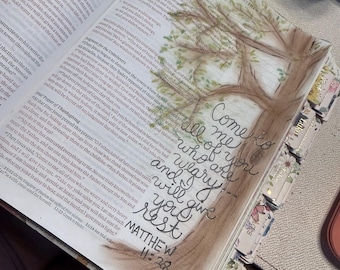 May include: An open Bible with printed text and a watercolor illustration of a tree. The handwritten calligraphy in black ink reads, Come to me all of you who are weary and I will give you rest, followed by the scripture reference Matthew 11:28.