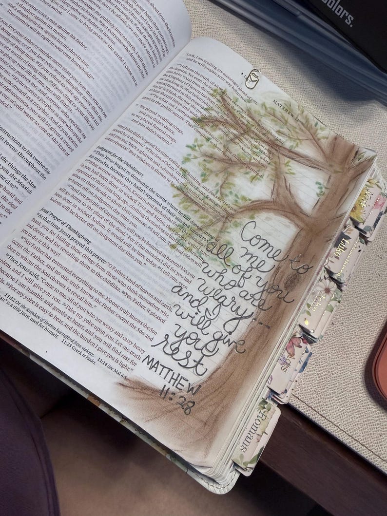Puede incluir: Una Biblia abierta con texto impreso y una ilustraci&oacute;n en acuarela de un &aacute;rbol. La caligraf&iacute;a escrita a mano en tinta negra dice, Come to me all of you who are weary and I will give you rest, seguida de la referencia b&iacute;blica Matthew 11:28.